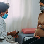 A pregnant woman speak with her doctor at a doctor's office, both are wearing masks.